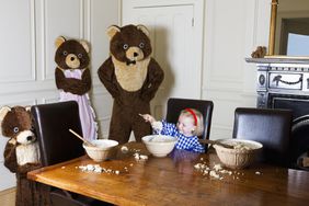A little girl eating from a bowl at a table surrounded by three bears