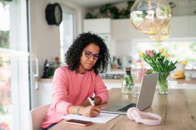 Bespectacled woman writing something on a paper while working on a laptop