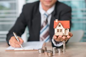 man in a suit and tie filling out paperwork holding a house figurine and house keys with pile of change on the table