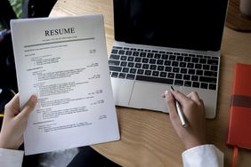 Image shows hands holding a multiple page resume, with a pen in one hand in front of a laptop.