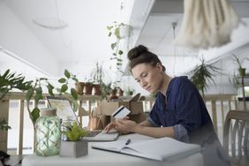 A young woman plant-shop owner sits at a light filled desk and scrutinizes her debit card.