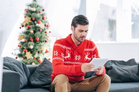 person in red and white holiday sweater sitting with tablet in front of christmas tree