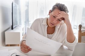 Woman looking worried holding paperwork at home. She is reading a financial bill or a letter with bad news. She looks very stressed and upset. There is a laptop computer on the table.