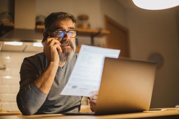 Man on phone with paperwork in front of laptop