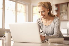 Woman in kitchen smiles while typing on a laptop