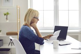 Blond woman in blue abrash sweater reviews papers at a desk near a laptop