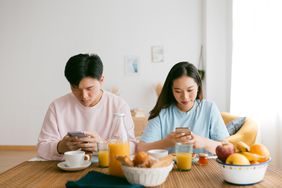 A couple sits next to each other on their phones, not paying attention to each other. Breakfast is on the table in front of them. 