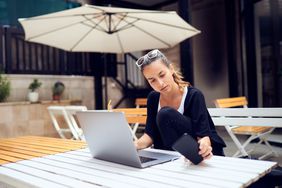 Image shows a woman sitting on a veranda working on her laptop.