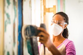 A woman wears a facemask and goggles while sanding a wall with an electric sander