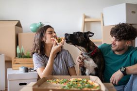 A young couple and their dog take a break from unpacking moving boxes to eat pizza on the floor of their new home