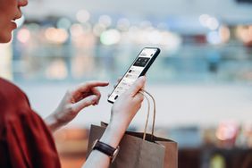 Cropped shot of young woman holding a paper shopping bag, managing online banking with mobile app on smartphone