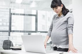 Pregnant woman standing at her desk reading over her client maternity email before she sends it.