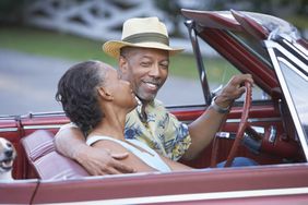 A man in a straw fedora looks adoringly at his wife as they drive a convertible