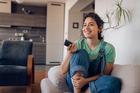 A woman smiling and sitting on a couch in a living room holding up a tv remote