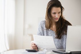 Brunette woman with calculator holds paperwork while looking at laptop