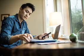 Seated man adds up Roth IRA savings with calculator and laptop at home
