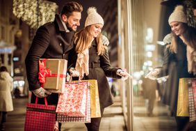 A couple holding shopping bags walks along the sidewalk looking in windows at holiday items.