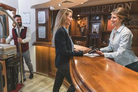 Woman paying with her credit card in a hotel; front desk receptionist is smiling, and bellhop is waiting with her luggage 