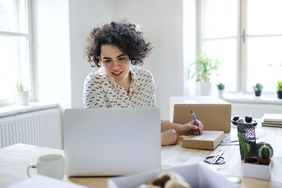 Woman sits in home office with laptop writing on a package, getting ready to mail it