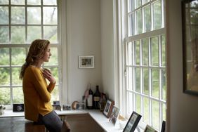 Woman with a coffee mug looking thoughtfully outside window of family home