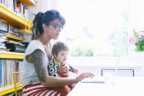Woman with sleeve tattoo uses laptop with baby in lap