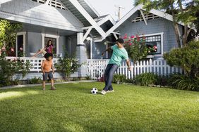 A man kicks a soccer ball to a youth in front of a home. 