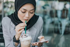 Woman drinking frothy coffee while working on smartphone