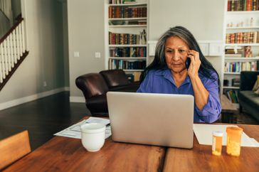 A woman sits at her kitchen table with laptop while paying bills, talking on the phone 
