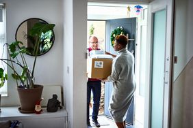 A delivery man hands a box of merchandise to a woman at her front door