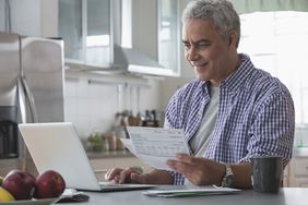 Grey haired man working on laptop with paper financial statements in one hand