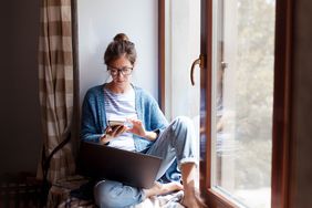 Woman working on laptop and phone near window at home