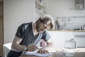 A man sits at home writing on a document with a glass of water in front of him