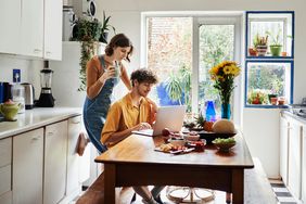 two people sitting in light kitchen with food on the counter, reading something on a computer