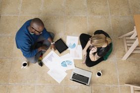Man and woman sitting on floor of home doing work while drinking coffee