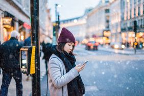 Woman stands on city corner with a hat on looking at a cell phone in snowy weather