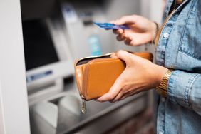 close up of a person holding a wallet in their left hand and an atm card in their right, in front of an atm