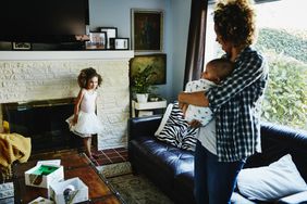 A parent holds a baby as a toddler plays in a home^as living room.