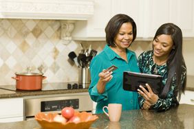 Mother and daughter buy something together in kitchen from an tablet while the mother holds a credit card.