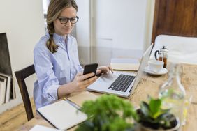 Woman using cell phone and laptop on wooden desk at home