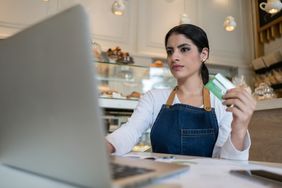 A bakery owner holds a business credit card in her hand as she prepares to place an order on her laptop