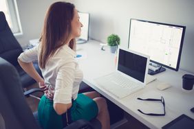 Woman sitting at desk in office chair holding her lower back in pain