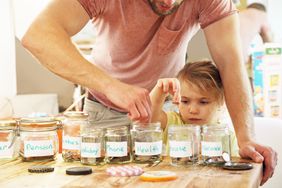 A child and parent drop coins into jars labeled with household expenses.