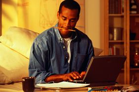 Man at Home Laptop Reading Papers and Typing With Pencil in His Mouth