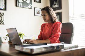 Woman in red shirt sitting at a desk in a home office typing on a laptop.
