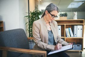 Woman in blazer and glasses filling out paperwork on clipboard in office