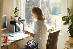 Woman drinking coffee and reading paperwork at desk in sunny home office