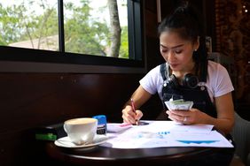 Female business owner counts money at a table with pen and paper while drinking a coffee