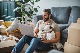 Man using laptop surrounded by moving boxes with dog nearby