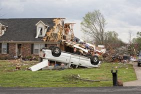A smashed and overturned SUV lies on the lawn in front of a wind-damaged home.