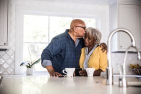 An older man kisses an older woman on the head while standing in the kitchen with coffee mugs on the counter